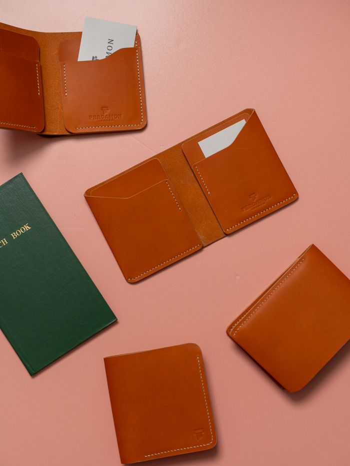 Brown leather wallets displayed on a pink surface alongside a green notebook, highlighting minimalist design.